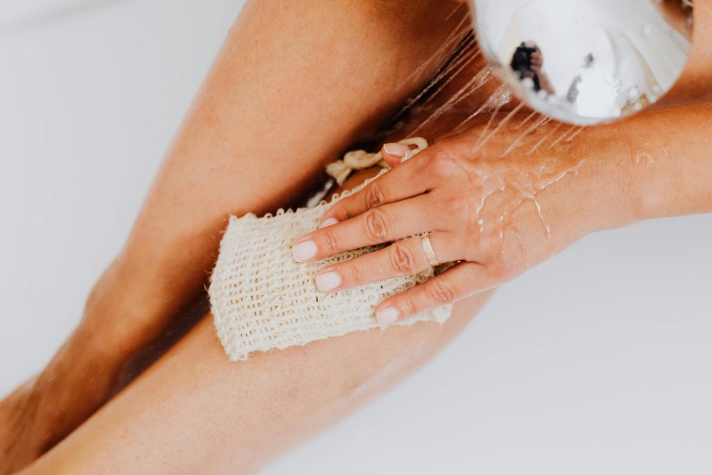 Close-up of a person exfoliating legs in the shower with a body scrubber.